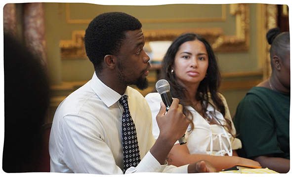 a young man speaks into a microphone at a town-hall-style public event