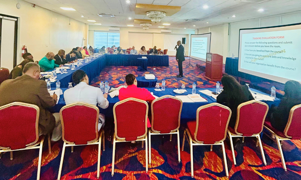 Dr Roger Koranteng stands and speaks into a microphone while giving training to participants sitting around a table in Trinidad and Tobago